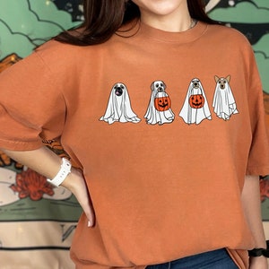 May include: A burnt orange t-shirt with a graphic of four dogs dressed as ghosts for Halloween. The dogs are wearing white ghost costumes with black eyes and mouths. One dog is holding a pumpkin bucket.