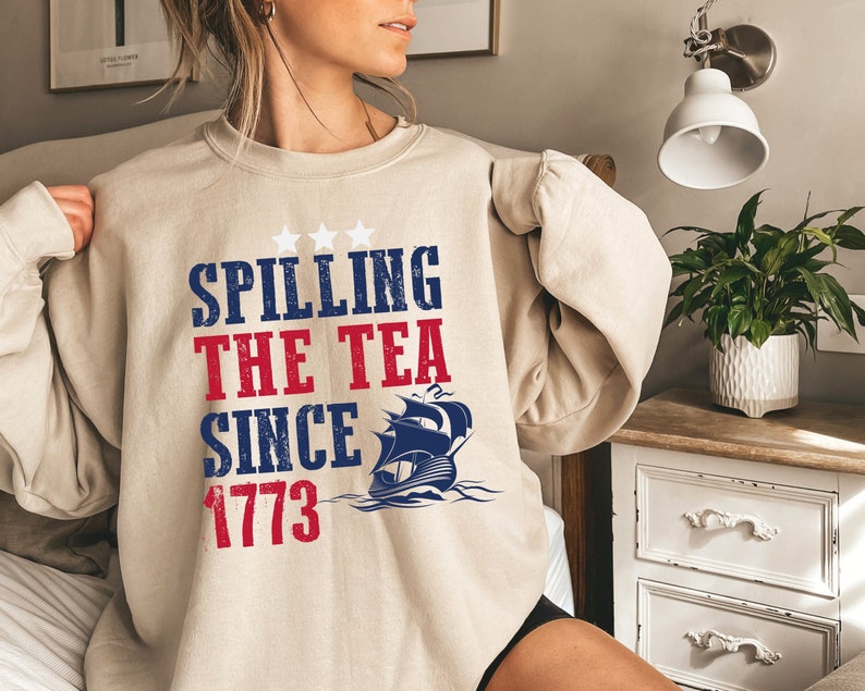 May include: A beige sweatshirt with the text "Spilling the Tea Since 1773" in red, white, and blue. A blue and white ship with three stars is above the text.