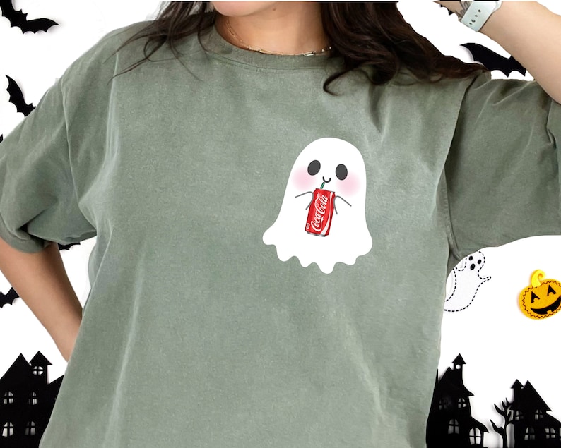 May include: A green t-shirt with a white ghost wearing a red Coca-Cola can. The ghost has a happy face and is holding the can with its hands.