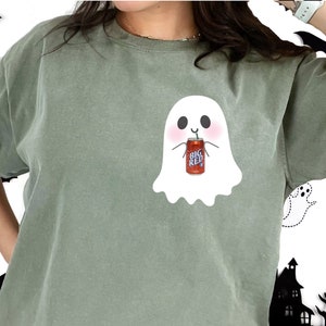 May include: A green t-shirt with a white ghost holding a can of Big Red soda. The ghost has a smiling face and is wearing a straw.