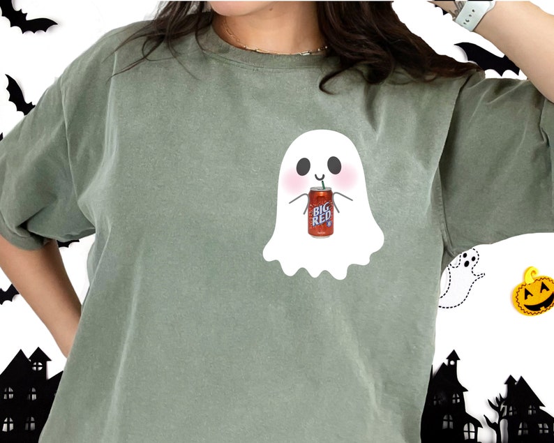 May include: A green t-shirt with a white ghost holding a can of Big Red soda. The ghost has a smiling face and is wearing a straw.