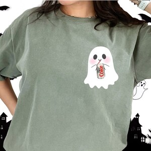 May include: A green t-shirt with a white ghost design holding a can of Diet Coke. The ghost has a cute face and is drinking from a straw.