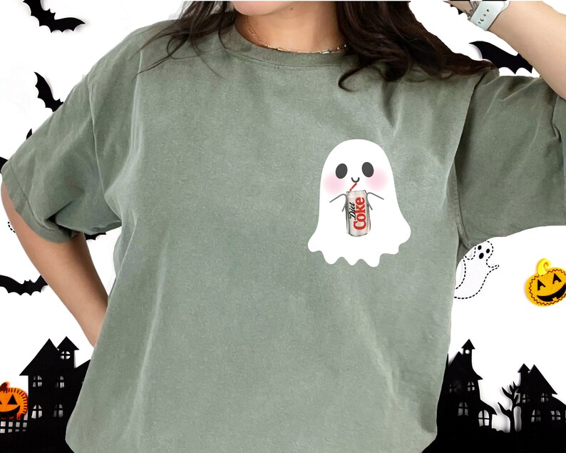 May include: A green t-shirt with a white ghost design holding a can of Diet Coke. The ghost has a cute face and is drinking from a straw.
