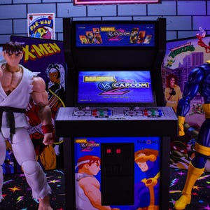 May include: Action figures of Ryu from Street Fighter and Cyclops from X-Men stand near a miniature arcade cabinet. The arcade cabinet has the game title "Marvel vs. Capcom" on the screen and side panels. Background includes X-Men and Teenage Mutant Ninja Turtles posters.