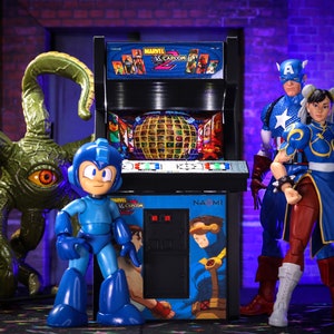 May include: A miniature arcade cabinet with a blue and black design. The cabinet features the text "MARVEL VS. CAPCOM 2" and "NAOMI".  The cabinet is surrounded by action figures, including a blue robot, a red, white, and blue superhero, and a female character in a blue and red outfit.
