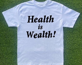 Puede incluir: Camiseta blanca con la frase "Health is Wealth!" impresa en letras negras en negrita. La camiseta tiene cuello redondo y mangas cortas. La prenda está hecha de un material suave y se muestra sobre una superficie verde y herbosa.