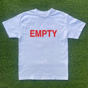 May include: White t-shirt with the word "EMPTY" printed in red block letters across the chest. The shirt is made of a soft material and has short sleeves and a crew neck. The shirt is displayed on a green surface.