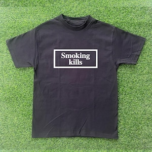 Könnte beinhalten: Schwarzes T-Shirt mit dem Text "Smoking kills" in einem weißen Rechteck. Das T-Shirt hat kurze Ärmel und einen Rundhalsausschnitt. Das Kleidungsstück wird auf einer grünen, grasbewachsenen Oberfläche präsentiert.