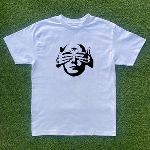 May include: White t-shirt featuring a black graphic of a face with hands covering the eyes and a single eye on the forehead. The shirt is made of a soft material and has short sleeves.
