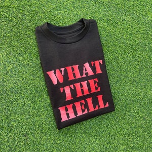 Op de afbeelding: Zwart T-shirt met de tekst "WHAT THE HELL" in rood gedrukt op de voorkant.