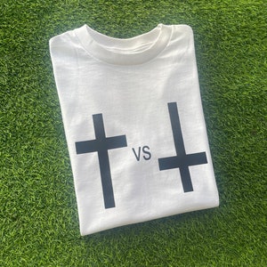 May include: White t-shirt with a black cross and an upside-down black cross printed on the front. The text "VS" is printed between the crosses.