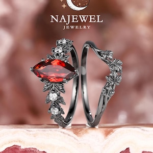 Könnte beinhalten: Zweiteiliges schwarzes Ring-Set. Ein Ring hat einen rotfarbenen, marquiseförmigen Edelstein und weiße Akzentsteine mit floralem Design. Der zweite Ring hat eine geschwungene Form mit passendem floralem Design. Die Ringe werden auf einer rosa Oberfläche präsentiert. Der Markenname "NAJEWEL JEWELRY" ist sichtbar.