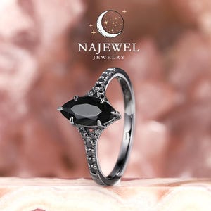 May include: A silver ring with a marquise-cut black gemstone. The ring features smaller black stones along the band and around the main stone. The ring is set against a pink and white background, with the text "NAJEWEL JEWELRY" in the upper left corner.