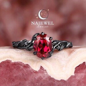 Può includere: Anello nero con una pietra preziosa ovale rossa incastonata in un design a foglia. L'anello è su uno sfondo rosa e bianco. NAJEWEL JEWELRY è scritto sopra l'anello.