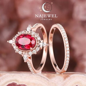 May include: Two rose gold rings. One ring features a large oval, red gemstone surrounded by smaller white gemstones. The second ring is a thin band with small, white gemstones. The rings are displayed on a pink surface. The brand name "NAJEWEL" is visible.