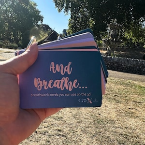 May include: A collection of teal, pink, and purple breathwork cards, featuring the words "And Breathe..." in a cursive script. The cards are designed for use on the go. The background shows a park with trees and a fountain.