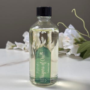 May include: A clear glass bottle with a black cap, containing a light yellow liquid. The bottle features a green label with the text "Whispers of Oudh" and "Cashmere Oudh". White flowers and a neutral grey backdrop are in the background.