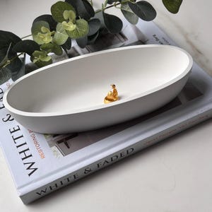 May include: A white oval ceramic dish with a gold-coloured ring on the inside. The dish is sitting on a book with the text "White & Faded" on the cover.