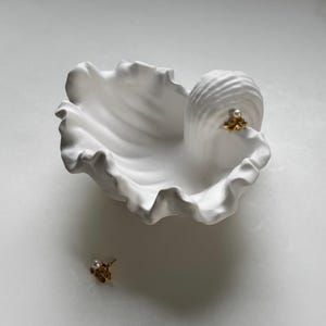 May include: A white, ceramic seashell-shaped dish with a ruffled edge. A small, gold-coloured earring with a pearl is placed on the dish, and another earring is next to the dish. The earrings have a floral design.
