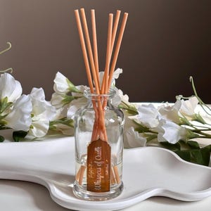 May include: A clear glass diffuser bottle with orange reeds. The bottle has a brown label that reads "Whispers of Oudh". The diffuser sits on a white, wavy tray, with white flowers in the background.