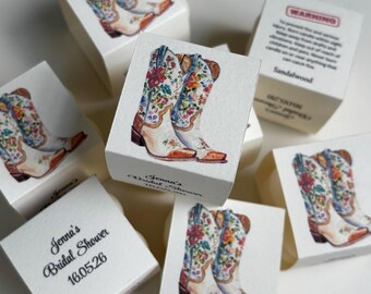 Könnte beinhalten: Weiße quadratische Kerzenschachteln mit einer Aquarellillustration eines Paares Cowboystiefel mit bunten Blumenmustern. Die Schachteln sind mit dem Text Jenna's Bridal Shower 16.05.26 bedruckt und enthalten ein Warnetikett an der Seite.