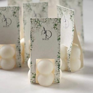 May include: Wedding favours in a cream colour, each with a double-sphere design, presented in decorative card holders. The cards feature floral accents, the initials "AD", and the date "30.11.25". The favours are on a light surface.