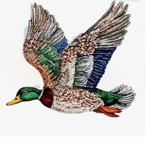 Könnte beinhalten: Ein fliegendes, mit Juwelen besetztes Enten-Design mit einer lebendigen Farbpalette. Die Flügel zeigen Brauntöne, Blau, Grün und Weiß, während der Körper Grün, Rot und Weiß aufweist. Die Ente hat einen gelben Schnabel und ist vor weißem Hintergrund.