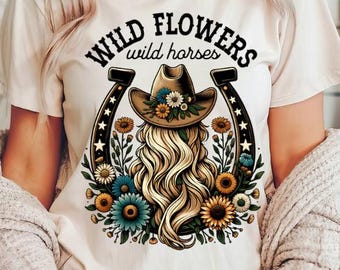 Puede incluir: Una camiseta blanca con un gráfico de una herradura con el pelo de una mujer y un sombrero de vaquero dentro. El texto "Wild Flowers wild horses" está por encima de la herradura.