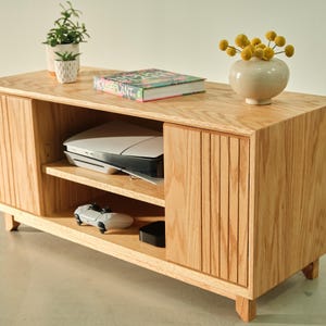 Puede incluir: Un mueble de TV de madera clara con puertas correderas y dise&ntilde;o de listones. La unidad tiene estantes abiertos con una consola de juegos, un controlador y otros aparatos electr&oacute;nicos. Un peque&ntilde;o jarr&oacute;n con flores amarillas y un libro encima.