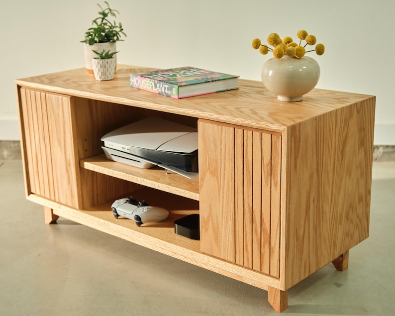 Puede incluir: Un mueble de TV de madera clara con puertas correderas y dise&ntilde;o de listones. La unidad tiene estantes abiertos con una consola de juegos, un controlador y otros aparatos electr&oacute;nicos. Un peque&ntilde;o jarr&oacute;n con flores amarillas y un libro encima.