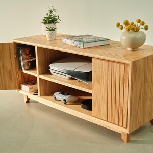 Puede incluir: Un mueble de entretenimiento de madera clara con estanter&iacute;as abiertas y una puerta de armario. El mueble contiene una impresora, una consola de juegos y otros art&iacute;culos. Una peque&ntilde;a planta y un jarr&oacute;n con flores amarillas encima.