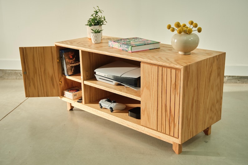 Puede incluir: Un mueble de entretenimiento de madera clara con estanter&iacute;as abiertas y una puerta de armario. El mueble contiene una impresora, una consola de juegos y otros art&iacute;culos. Una peque&ntilde;a planta y un jarr&oacute;n con flores amarillas encima.