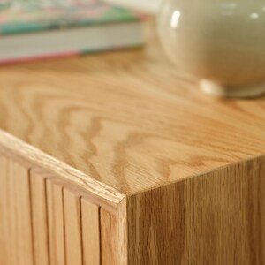 Puede incluir: Primer plano de un mueble de madera clara con un acabado de vetas naturales. Un jarr&oacute;n decorativo se encuentra en la parte superior, con un libro visible en el fondo. El mueble tiene un dise&ntilde;o de listones verticales en el lateral.