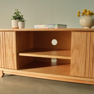 Puede incluir: Un mueble de TV de madera clara con dos puertas y un estante central abierto. Las puertas tienen ranuras verticales. Un peque&ntilde;o jarr&oacute;n con flores amarillas, un libro y plantas en macetas est&aacute;n encima. El mueble mide unos 152 cm de ancho.