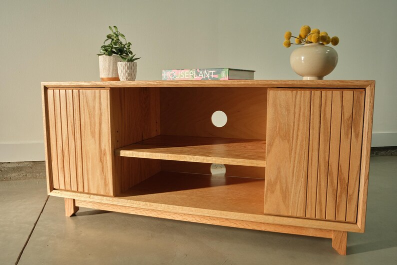 Puede incluir: Un mueble de TV de madera clara con dos puertas y un estante central abierto. Las puertas tienen ranuras verticales. Un peque&ntilde;o jarr&oacute;n con flores amarillas, un libro y plantas en macetas est&aacute;n encima. El mueble mide unos 152 cm de ancho.