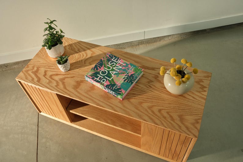 Puede incluir: Una mesa de centro de madera clara con un acabado de vetas de madera natural. La mesa presenta un libro titulado "HOUSE PLANT" con una portada colorida, dos peque&ntilde;as plantas en macetas y un jarr&oacute;n blanco con flores amarillas. La mesa tiene un estante.