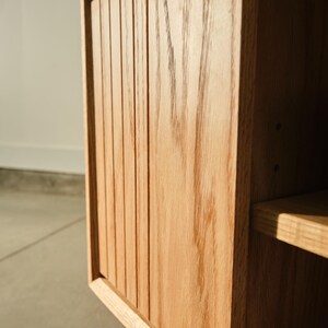 Puede incluir: Primer plano de un mueble de madera con acabado marr&oacute;n claro. La puerta del mueble presenta detalles verticales ranurados. Se ve una balda en el lado derecho. El mueble es de madera maciza y tiene veta natural.