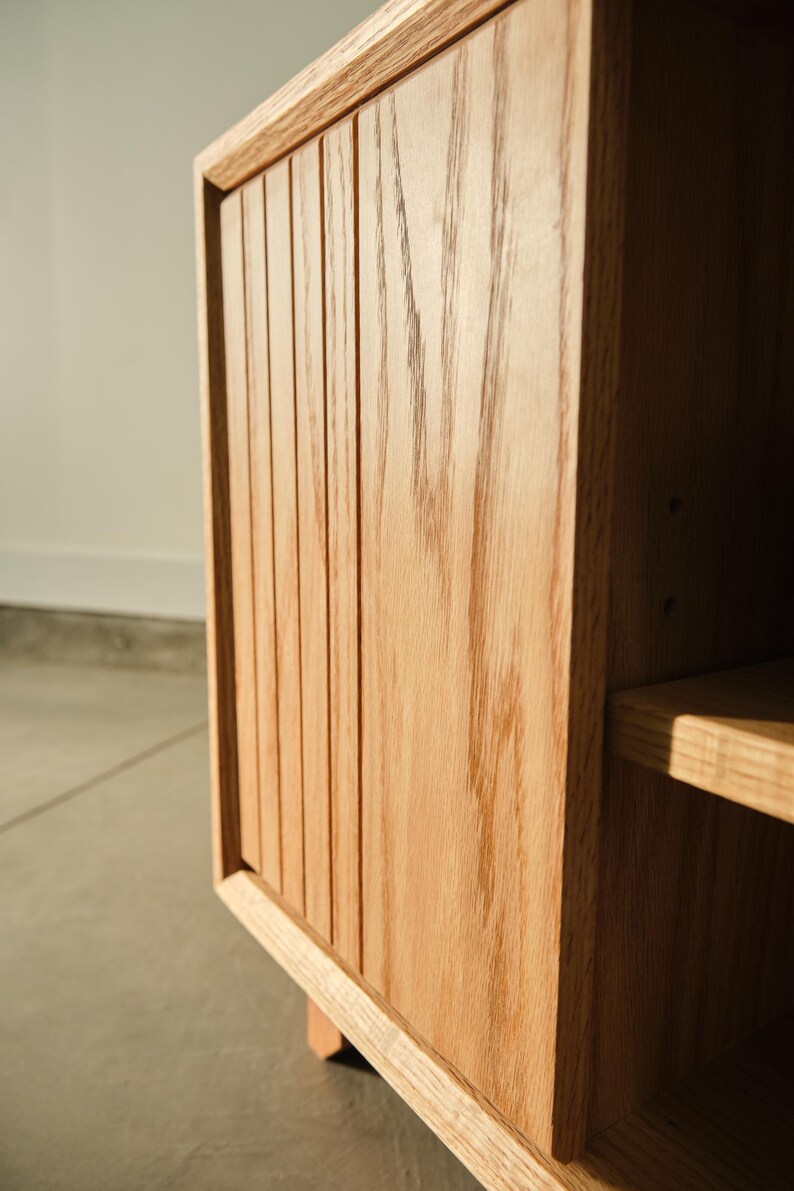 Puede incluir: Primer plano de un mueble de madera con acabado marr&oacute;n claro. La puerta del mueble presenta detalles verticales ranurados. Se ve una balda en el lado derecho. El mueble es de madera maciza y tiene veta natural.