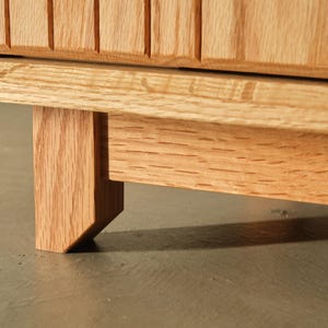 Puede incluir: Primer plano de un mueble de madera con acabado natural. El mueble presenta un dise&ntilde;o acanalado en la puerta y una pata robusta e inclinada. La veta de la madera es visible, mostrando el color y la textura naturales del material.