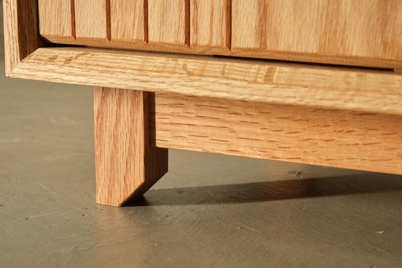 Puede incluir: Primer plano de un mueble de madera con acabado natural. El mueble presenta un dise&ntilde;o acanalado en la puerta y una pata robusta e inclinada. La veta de la madera es visible, mostrando el color y la textura naturales del material.