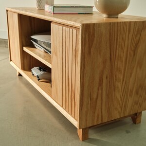 Puede incluir: Mueble de entretenimiento de madera clara con dos puertas correderas y estanter&iacute;as abiertas. La unidad tiene un acabado de veta de madera natural y est&aacute; soportada por patas cortas. Una consola de juegos y un mando son visibles en el interior.