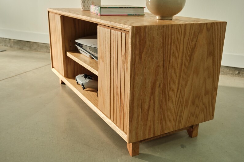 Puede incluir: Mueble de entretenimiento de madera clara con dos puertas correderas y estanter&iacute;as abiertas. La unidad tiene un acabado de veta de madera natural y est&aacute; soportada por patas cortas. Una consola de juegos y un mando son visibles en el interior.