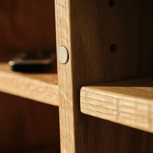 Puede incluir: Primer plano de una estanter&iacute;a de madera con acabado natural. La unidad presenta un patr&oacute;n de vetas visible y un peque&ntilde;o cierre magn&eacute;tico redondo de color plateado. Los estantes son horizontales y parecen resistentes.