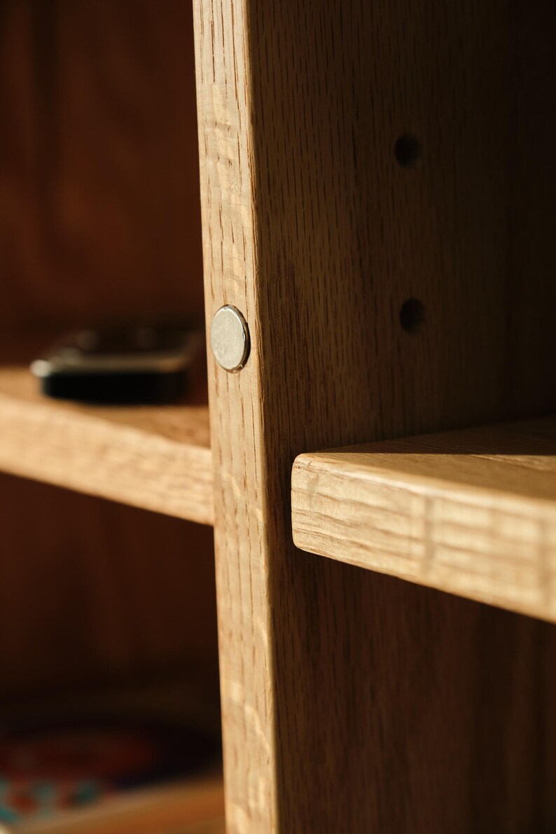 Puede incluir: Primer plano de una estanter&iacute;a de madera con acabado natural. La unidad presenta un patr&oacute;n de vetas visible y un peque&ntilde;o cierre magn&eacute;tico redondo de color plateado. Los estantes son horizontales y parecen resistentes.