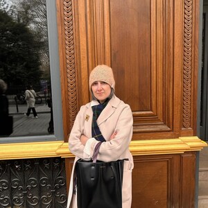 May include: A woman wearing a beige coat, a patterned scarf, and a knit hat, holding a large black tote bag. The background features a wooden door and a window with a decorative metal grate.
