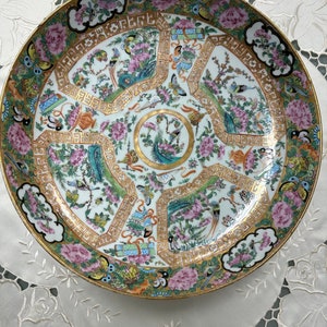 May include: A large, round porcelain plate with a white background and intricate gold and pink floral designs. The plate features a central motif of a peacock with its tail feathers spread out. The border of the plate is decorated with a repeating pattern of stylized flowers and leaves.