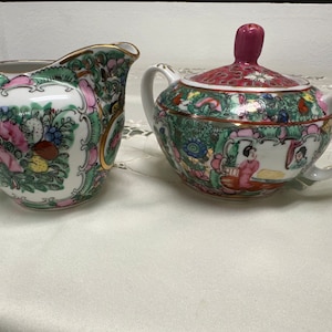 May include: A vintage porcelain creamer and sugar bowl set with a floral and figural design. The creamer and sugar bowl are both white with green, pink, and gold accents. The sugar bowl has a red lid with a floral design.