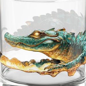 Könnte beinhalten: Ein klares Glas mit einer detaillierten Illustration eines grünen und goldenen Alligators. Das Kunstwerk umschließt das Glas und zeigt den Kopf und den Körper des Reptils. Das Design hat ein strukturiertes, künstlerisches Aussehen.