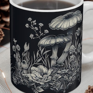 Könnte beinhalten: Eine schwarze Keramiktasse mit weißem Henkel zeigt eine detaillierte Illustration von Pilzen und Blumen im Vintage-Stil. Die Tasse ist mit einer dunklen Flüssigkeit gefüllt, wahrscheinlich Kaffee oder Tee.