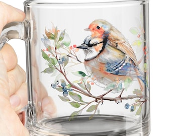 May include: A clear glass mug with a handle features a watercolor illustration of a bluebird perched on a branch with berries. The bird has a blue, white, and orange plumage.